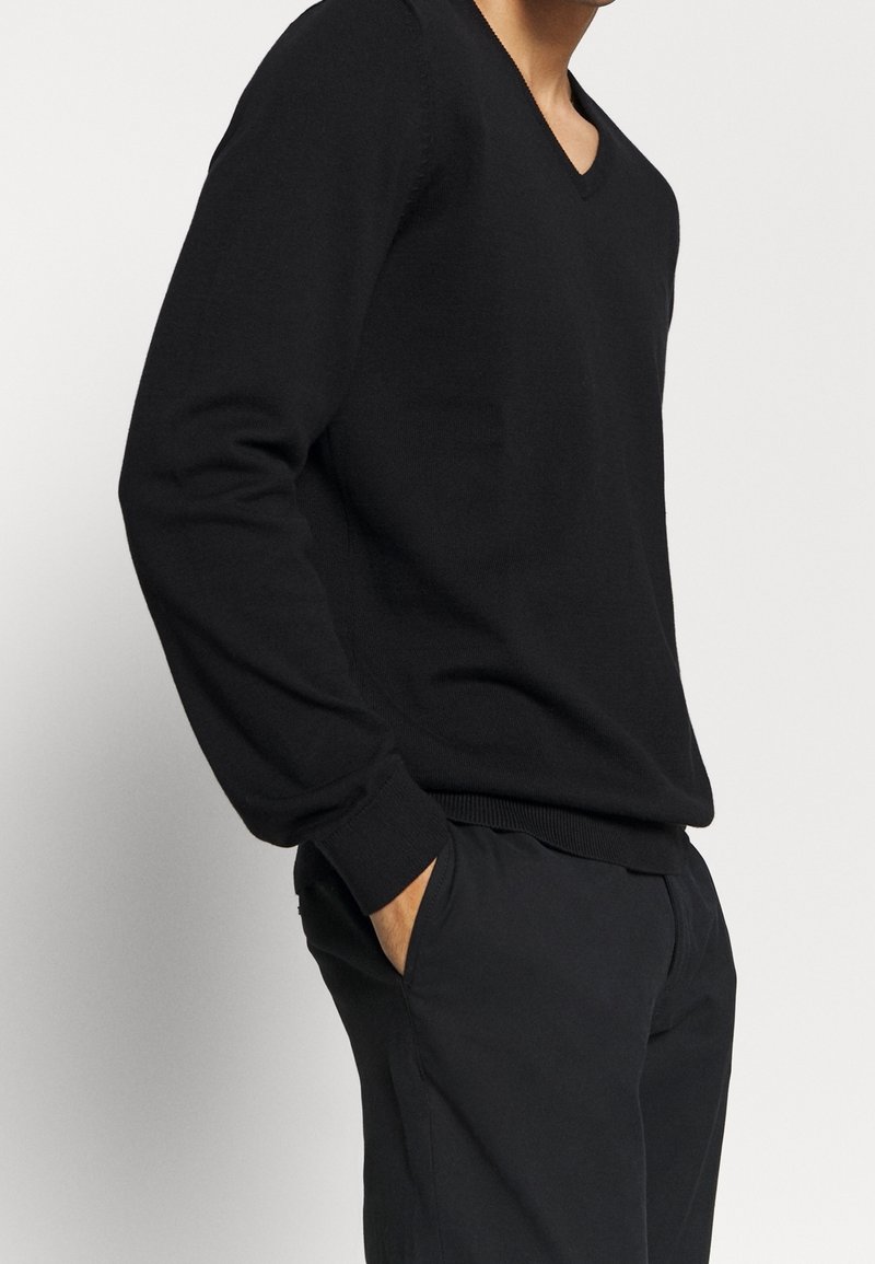 Maglione nero lavorato a maglia con scollo a V, maniche lunghe e polsini a coste. Indossato sopra pantaloni scuri, presenta un design semplice e classico.