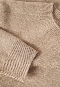 Pull beige tricoté avec une texture douce. Présente des poignets et un col côtelés, mettant en valeur un design simple et classique.