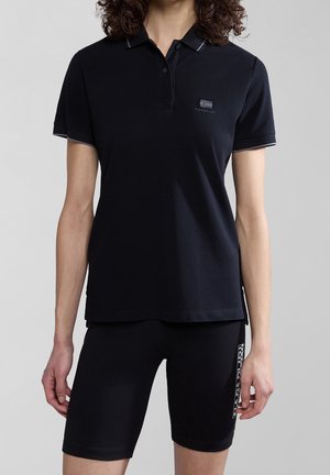 Person trägt ein schwarzes Polo-Shirt mit kurzen Ärmeln und schwarze eng anliegende Shorts mit Markenlogo auf Brust und Oberschenkel vor neutralem Hintergrund.
