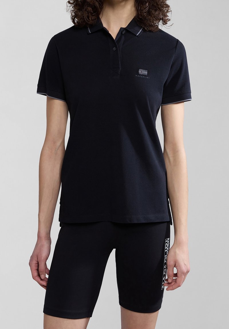 Personne portant un polo noir à manches courtes et un short noir ajusté avec un logo de la marque sur la poitrine et la cuisse, sur un fond neutre.
