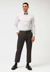 Weißes Hemd mit schwarzen Knöpfen und Manschettenknöpfen, kombiniert mit dunkler Hose und schwarzen Schuhen. Modell posiert selbstbewusst.