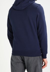 Felpa con cappuccio di color blu navy, dal texture morbido, con polsini e orlo a costine. Presenta una tasca a marsupio e una finitura liscia.