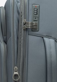 Valise grise avec une surface en tissu texturé, fermetures éclair à double sens et une serrure à combinaison intégrée. Présente un design élégant et moderne.