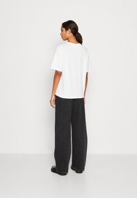 Witte T-shirt met korte mouwen en een relaxte pasvorm, gecombineerd met brede zwarte denim broek, getoond vanaf de achterkant tegen een lichte achtergrond.