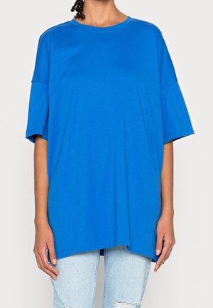 Personne portant un t-shirt ample à manches courtes bleu vif sur un jean déchiré bleu clair sur un fond uni.