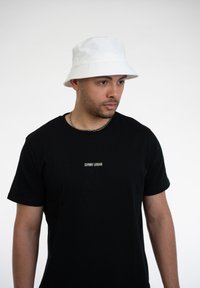 Biely klobúk s textúrovaným okrajom a jemným logom, nosený s čiernym tričkom s krátkym rukávom, ktoré má malý vyšívaný názov značky.