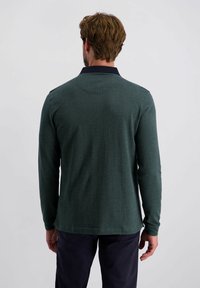 Donkergroene polo met lange mouwen, voorzien van een visgraatstructuur, marineblauwe kraag en zijsplitsen aan de zoom. De achterkant toont een getailleerd silhouet.