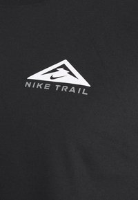 Černé bavlněné tričko s bílým trojúhelníkovým logem a textem "NIKE TRAIL" níže v moderním bezpatkovém písmu. Hladká textura.