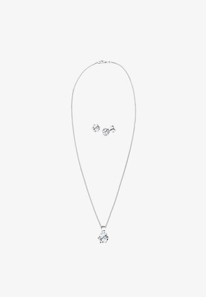 Ezüst nyaklánc lánccal és egy középső átlátszó medállal; a hozzáillő fültágítók átlátszó drágakövekből készült virágformákkal rendelkeznek.