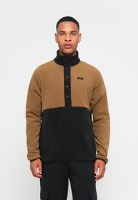 Pullover en polaire marron et noir avec un col haut, fermeture à boutons-pression et détail du logo sur la poitrine. Texture lisse et design sportif.