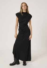 Abito maxi nero elasticizzato con maniche a cappuccio, design drappeggiato e dettagli arricciati in vita, abbinato a stivali neri alla caviglia.