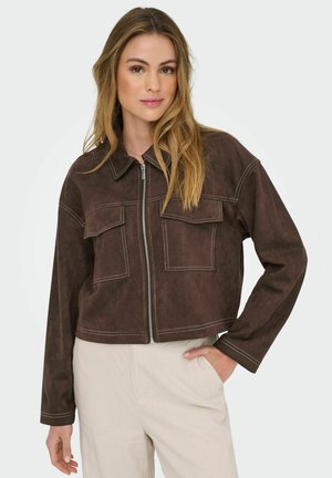 Kvinde med langt hår iført en brun cropped jakke med to brystlommer, parret med lyse bukser, stående mod en ensfarvet baggrund.