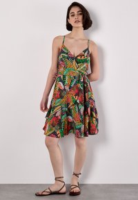 Junge Frau steht vor einem neutralen Hintergrund, trägt ein farbenfrohes, tropisches gemustertes Kleid und Sandalen mit Riemen und blickt selbstbewusst in die Kamera.