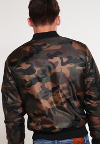 Kamuflāžas bumbiera jaka zaļos, brūnos un melnos toņos. Gluda materiāla, ribota apkakle un aproces, elastīga josta un bez redzamiem mehānismiem.