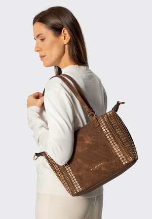 Frau in weißem Pullover, die eine braune Ledertasche mit goldenen Nieten und doppelten Schulterriemen trägt.