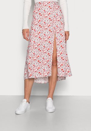 Jupe midi fleurie avec fente avant portée avec un haut blanc à manches longues et des baskets blanches, debout devant un fond uni.