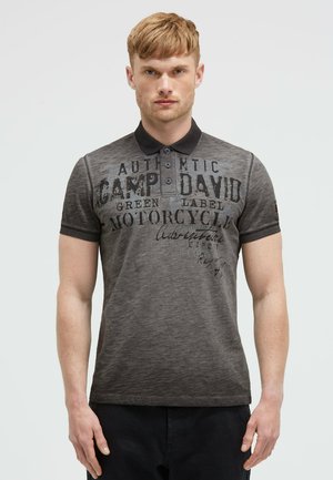 Junger Mann mit kurzen hellbraunen Haaren, der ein graues Poloshirt mit schwarzem Kragen und bedrucktem Text trägt, steht vor einem einfarbigen Hintergrund.