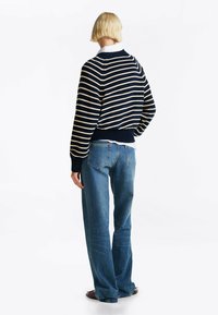 Maglione lavorato a maglia blu navy con strisce crema, polsini e orlo a costine. Abbinato a jeans larghi blu chiaro. Sfondo bianco uniforme.