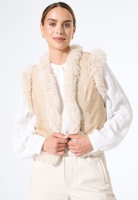 Vrouw in een beige nepleren vest met fake fur-bies over een witte blouse met lange mouwen en crèmekleurige broek, houdt de randen van het vest op taillehoogte vast.