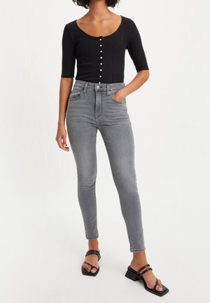 Femme portant un haut noir boutonné, un jean skinny gris taille haute et des sandales à talons noires à brides, debout avec une main dans la poche.