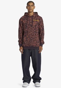 Sudadera con capucha de rayas color burdeos con cordón, que incluye un bolsillo frontal y un logo amarillo. Combinada con vaqueros oscuros holgados y zapatillas blancas.