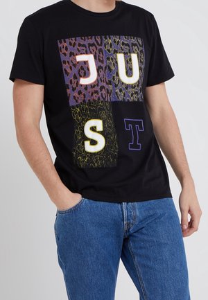 Mężczyzna ubrany w czarny t-shirt z wzorem w panterkę i literami J, U, S, T w kolorze białym i fioletowym, w połączeniu z niebieskimi dżinsami.