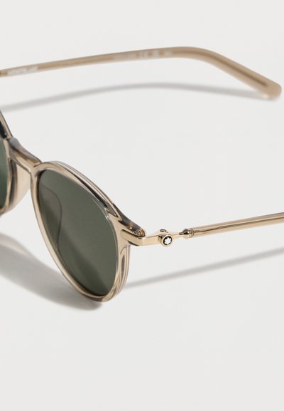 Στρογγυλά γυαλιά ηλίου με χρυσό μεταλλικό σκελετό και σκούρους πράσινους φακούς, εμφανίζονται σε κοντινή λήψη με λευκό φόντο.