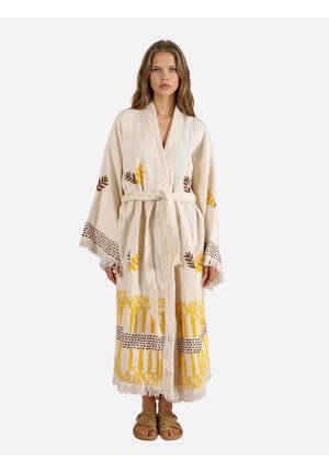 Kvinde stående, iført en beige badekåbe med gule og brune botaniske print, bundet om taljen, og beige sandaler med flettede remme.
