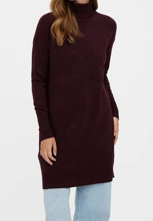 Femme portant un long pull à col roulé violet foncé associé à un jean bleu clair, debout devant un fond blanc uni.