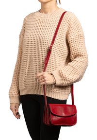 Borsa a tracolla rossa in pelle con chiusura a pattina, sottile tracolla e dettagli di cucitura visibili. Indossata con un maglione lavorato a maglia beige e pantaloni neri.