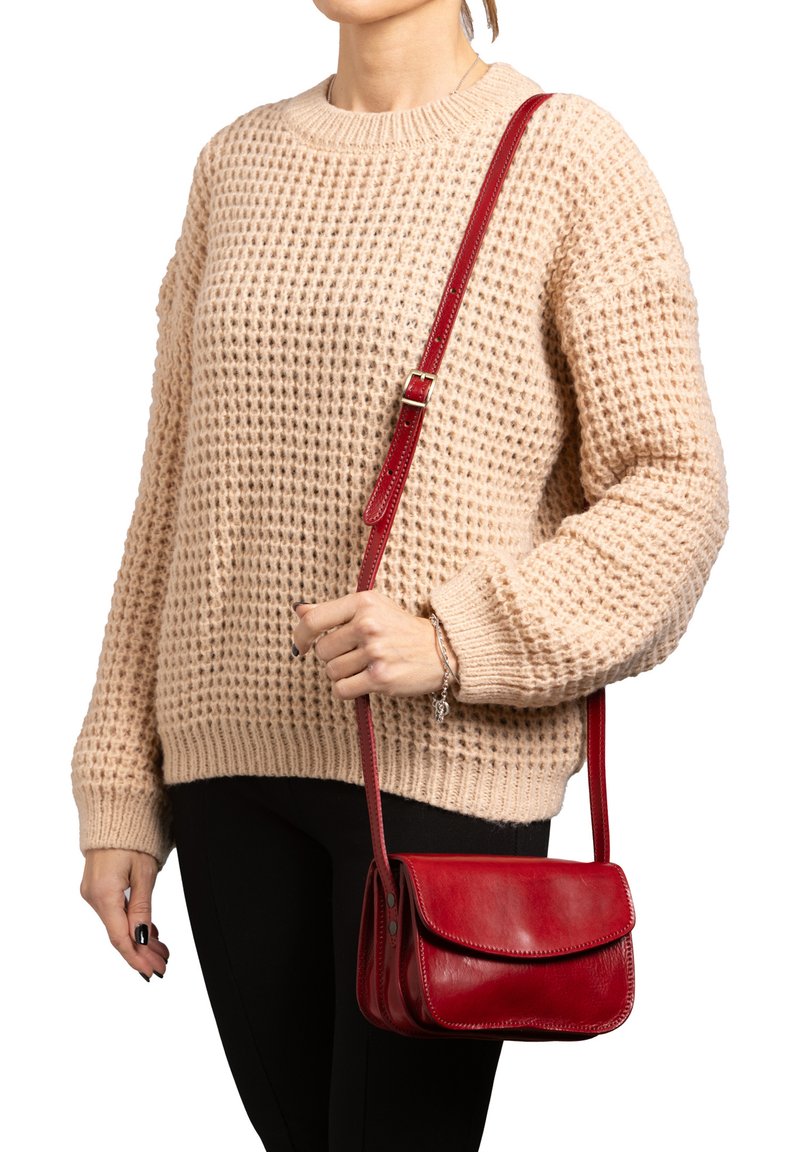Borsa a tracolla rossa in pelle con chiusura a pattina, sottile tracolla e dettagli di cucitura visibili. Indossata con un maglione lavorato a maglia beige e pantaloni neri.