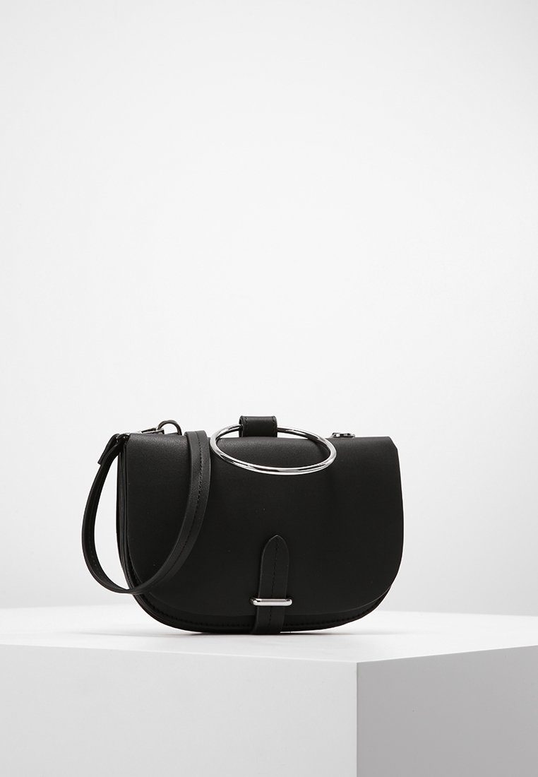 Zwarte leren schoudertas met zilveren ringhandgreep en gespdetails, tentoongesteld op een witte pedestaal tegen een effen achtergrond.