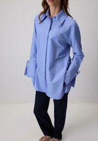 Plava i bijela prugasta oversized košulja s oštrim ovratnikom, dugmadima na rukavima i bočnim prorezima, u kombinaciji s tamnim hlačama i ležernim cipelama.