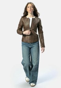 Bruine leren ritsjack met een getailleerd ontwerp en naadaccenten, gecombineerd met blauwe flared jeans en witte sneakers. Model staat rechtop.