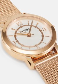 Roségoldene Mesh-Armbanduhr mit Ziffernblatt in Weiß, ausgestattet mit roségoldenen Stundenmarkierungen und Zeigern. In der Mitte befindet sich der Markenname "GUESS".