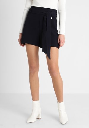 Slanke ben iført sorte shorts med høj talje og et stofbælte bundet om taljen, hvid ribbet langærmet top og hvide ankelstøvler med blokhæle.