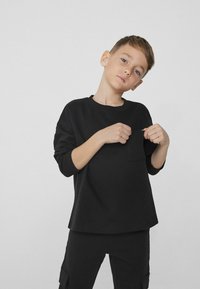 Czarna koszula z długim rękawem, okrągłym dekoltem i kieszenią z przodu, w połączeniu z dopasowanymi czarnymi spodniami, wykonanymi z miękkiego, gładkiego materiału.