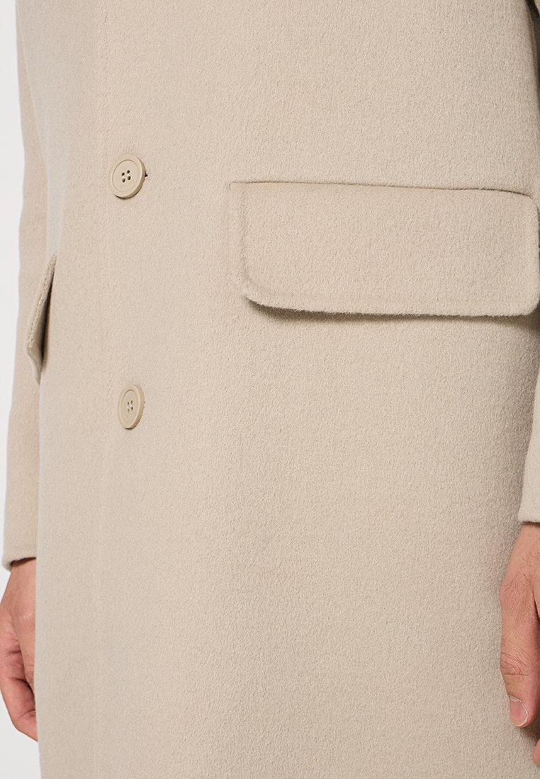 Beige Wollmantel mit texturiertem Finish, ausgestattet mit einer Zwei-Knopf-Verschluss und einer einzelnen Klappentasche auf der linken Seite.