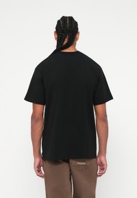 Black cotton t-shirt with a crew neckline, short sleeves, and a relaxed fit, paired with brown pants featuring a brand label on the pocket.