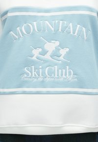 Helehall sinine pusa, millel on valge tikandiga tekst "MOUNTAIN Ski Club" ja suusatajate kujutised. Viimistletud valge triibuga.