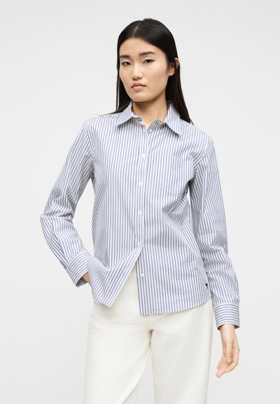 Νεαρή γυναίκα με μακριά σκούρα μαλλιά, ντυμένη με πουκάμισο με κάθετες ρίγες σε μπλε και άσπρο και άσπρο παντελόνι, στέκεται με το ένα χέρι στην τσέπη.