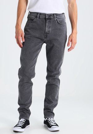 Man draagt slim-fit donkergrijze spijkerbroek, wit t-shirt en zwarte lage sneakers, staand tegen een effen lichte achtergrond.