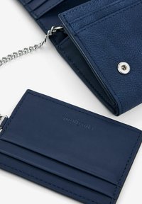 Navyblaues, strukturiertes Portemonnaie mit einer Kette, ausgestattet mit einem Druckverschluss und mehreren Kartenfächern. Enthält einen passenden Kartenhalter mit eingeprägtem Logo.