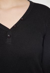 Černý ribbovaný pletený top s výstřihem do V, se třemi hnědými knoflíky na légě a jemnou texturou.