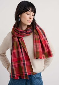 Rødt og grønt ternet tørklæde med en kombination af frynser og forskellige stribebredder, båret draperet over en lysefarvet sweater.