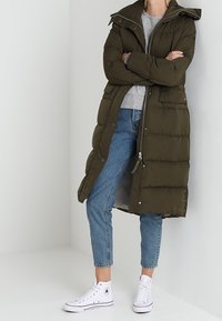 Mørkegrøn, knælang pufferjakke med lynlås og trykknaplukning, med hætte, oversized design og sidelommer. Bæres over en grå sweater og blå jeans.