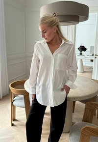 Fehér gombos ing, egyetlen zsebbel, hosszú ujjakkal és ívelt heméssel, fekete nadrággal viselve. Simább anyag, laza stílus.