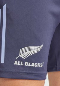 Tummansininen kangashousut, joissa on valkoinen saniaisen logo ja "ALL BLACKS" -teksti, suunniteltu urheiluun, joissa on sileä pinta ja moderni leikkaus.