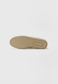 Beige skosåle med et mønstret gummibunn, som har en flat rektangulær aksent og en teksturert kant for bedre grep.