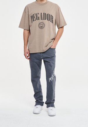 Beiges kurzärmeliges T-Shirt mit dem Schriftzug "PEGADOR", kombiniert mit dunkelgrauen Jeans mit weißem Signaturdetail. Weiße Sneaker vervollständigen das Outfit.